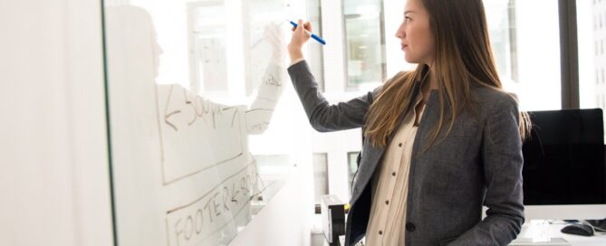 A woman wearing a gray blazer and writing on a whiteboard.