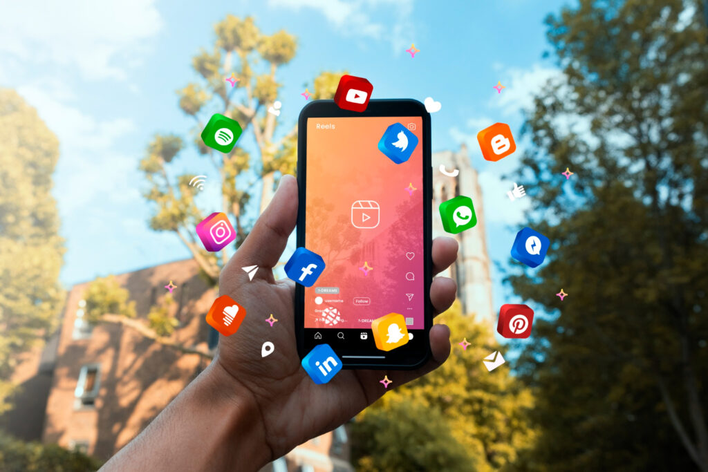 A hand holding a smartphone with various social media app icons floating around it 