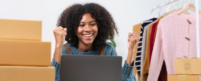 A smiling small business owner celebrates positive online results while working on a laptop in a home workspace surrounded by shipping boxes and products ready for customers.