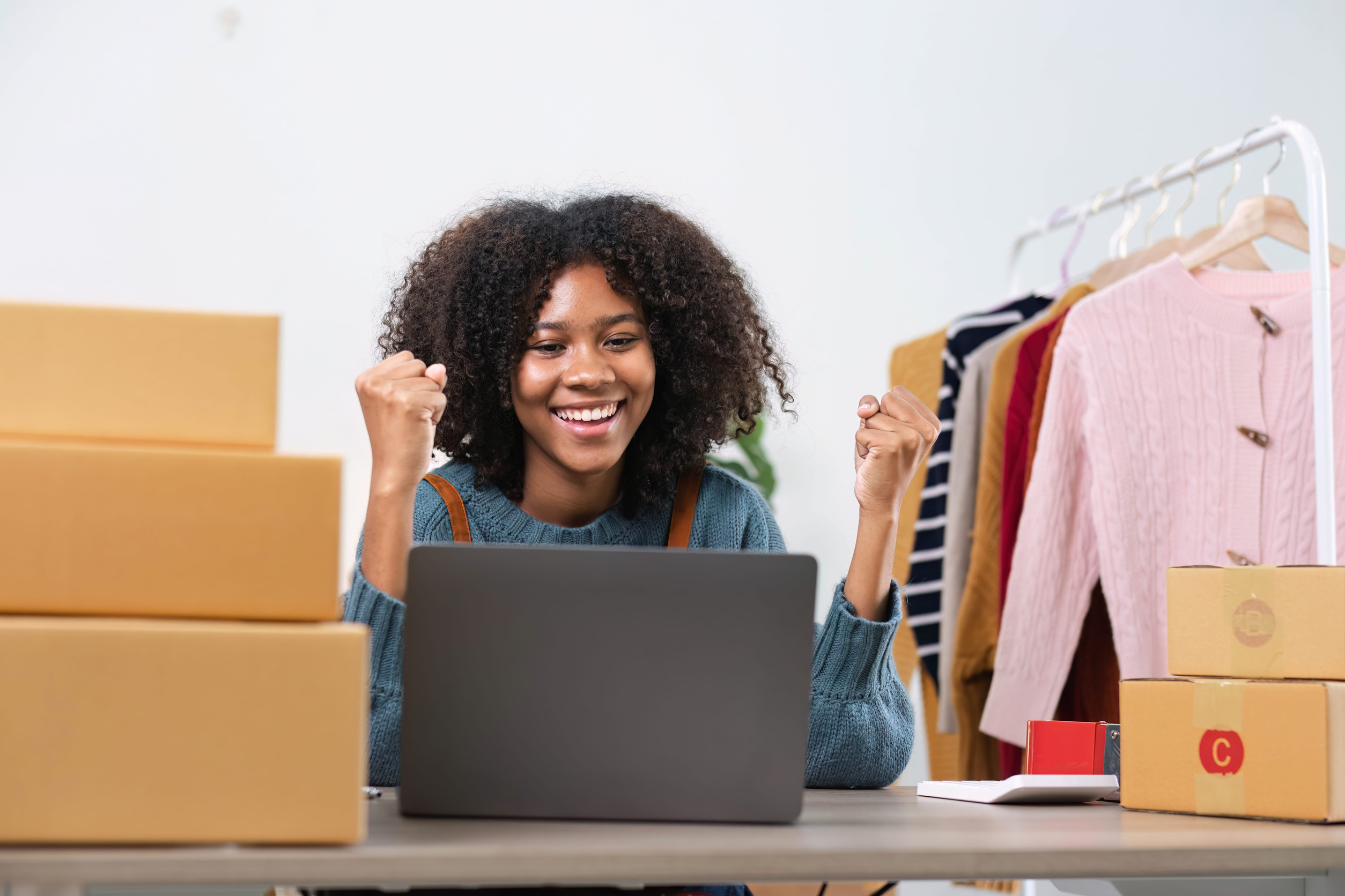 A smiling small business owner celebrates positive online results while working on a laptop in a home workspace surrounded by shipping boxes and products ready for customers.
