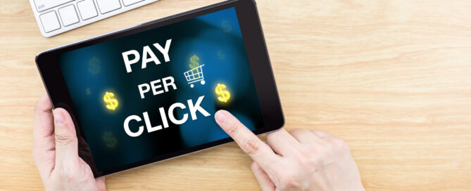 A person holds a tablet displaying the words "Pay Per Click" with dollar signs and a shopping cart icon, illustrating PPC digital advertising and paid online marketing.