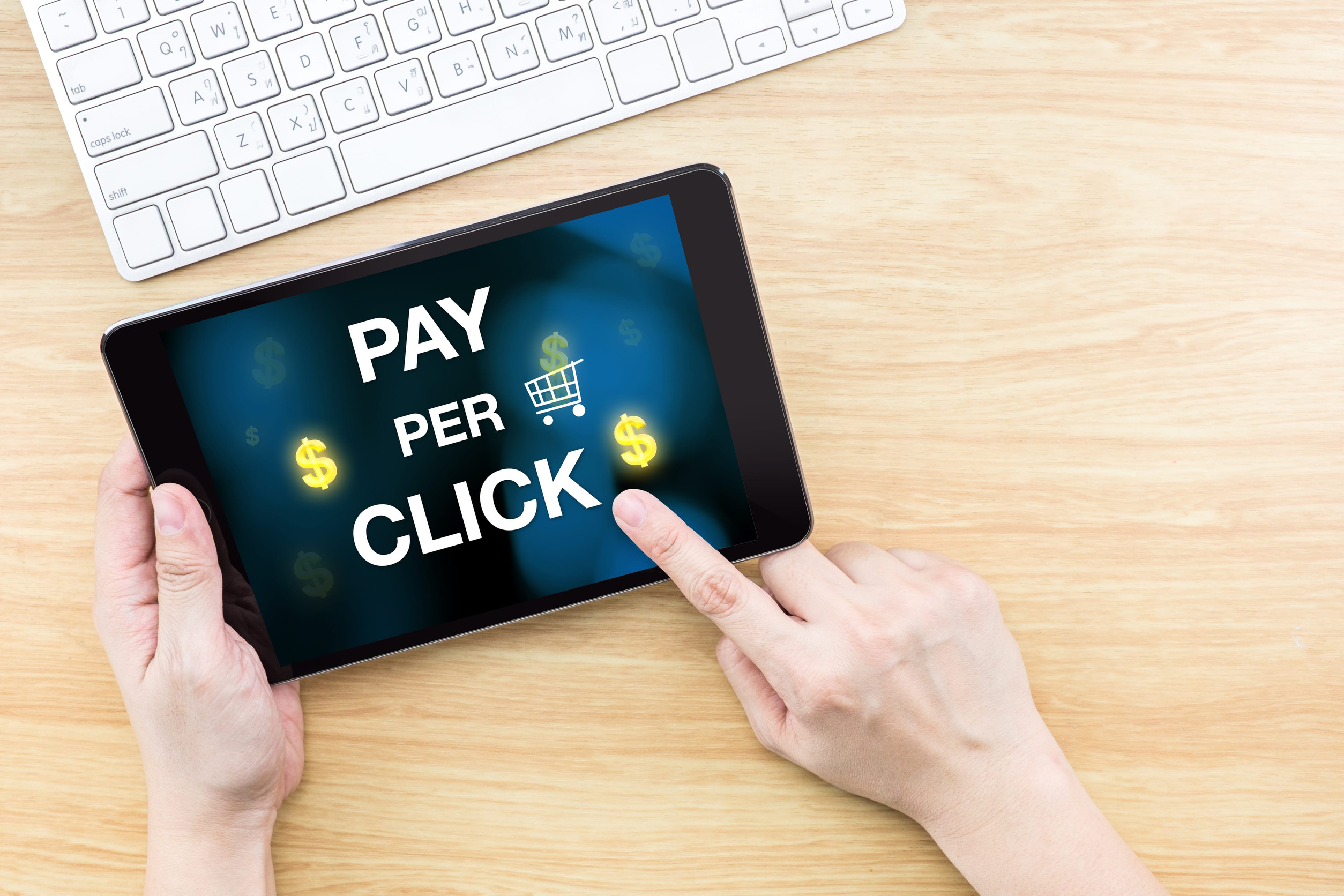 41205183_m A person holds a tablet displaying the words "Pay Per Click" with dollar signs and a shopping cart icon, illustrating PPC digital advertising and paid online marketing.
