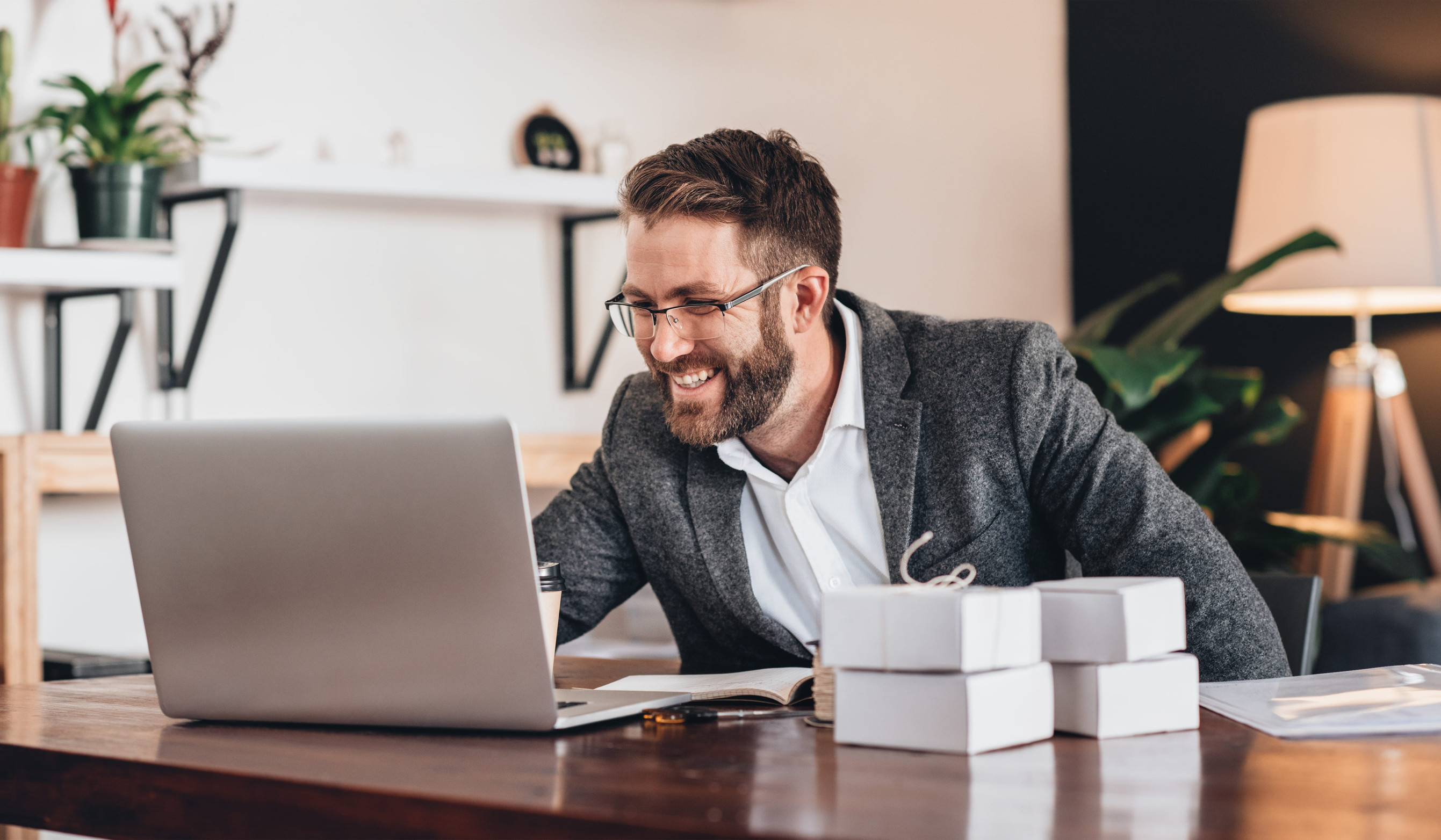 A smiling small business owner works on a laptop at a desk with product boxes nearby, representing online business growth through digital marketing and PPC advertising